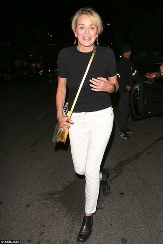 Sharon Stone wearing Gold Leather Crossbody Bag, Black Leather Chelsea Boots, White Jeans, Black Short Sleeve Sweater